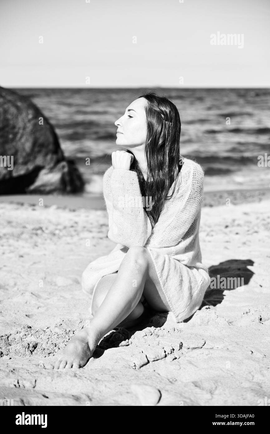 Junge Frau mit langen Haaren sitzt barfuß am Sandstrand an der Ostsee, mit geschlossenen Augen, in ruhigem Sonnenlicht mit heiterem Ausdruck Stockfoto