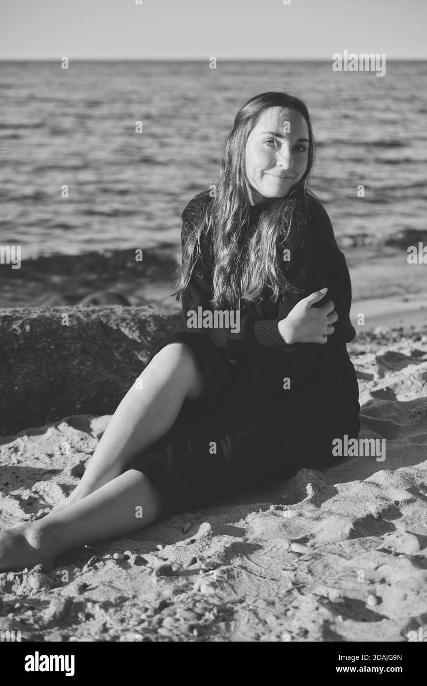 Porträt einer jungen Frau mit langen Haaren, die am Sandstrand an der Ostsee sitzt. Sanfte Beleuchtung sorgt für einen ruhigen Ausdruck und eine ruhige Küstenatmosphäre Stockfoto