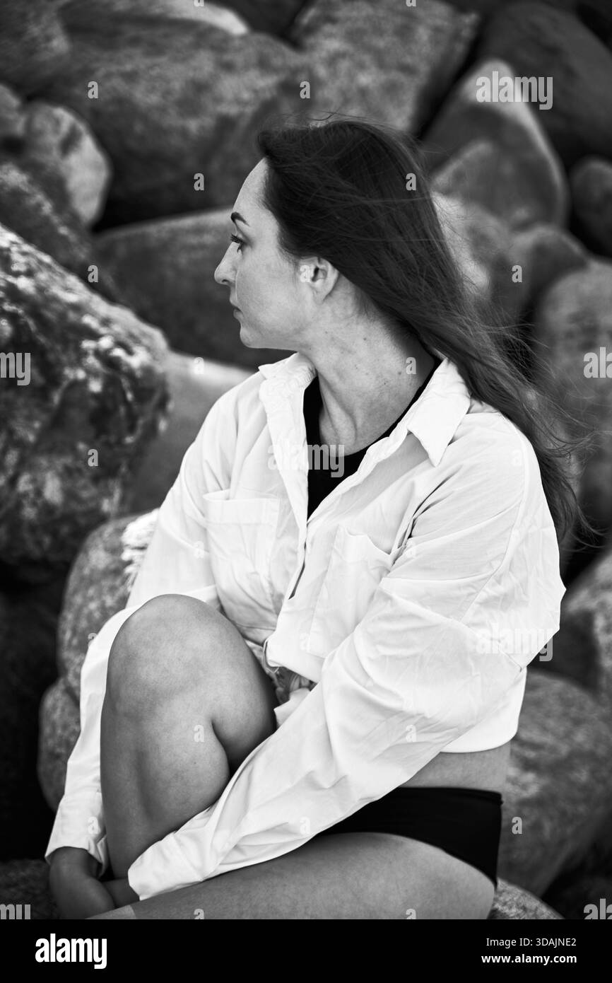 Junge Frau mit langen dunklen Haaren und weißem Hemd sitzt auf felsigem Gelände und blickt mit kontemplativem Ausdruck zur Seite. Natürliche Außenbeleuchtung hervorgehoben Stockfoto