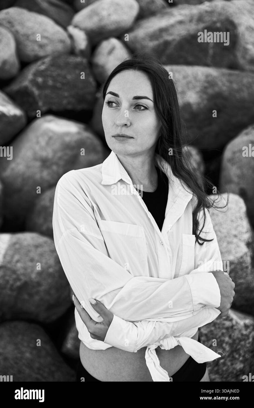 Junge Frau mit langen dunklen Haaren steht mit überkreuzten Armen vor großen Felsen und zeigt kontemplativen Ausdruck in sanftem natürlichem Licht. Monochrom Stockfoto