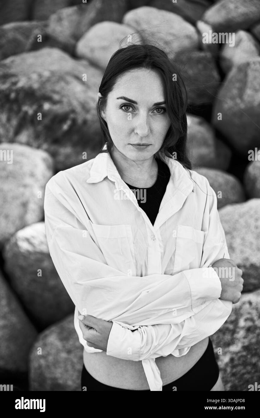 Schwarz-weißes Porträt einer jungen Frau mit dunklem Haar und intensivem Blick, die mit überkreuzten Armen vor großen Felsen steht. Natürliche Umgebung im Freien A Stockfoto