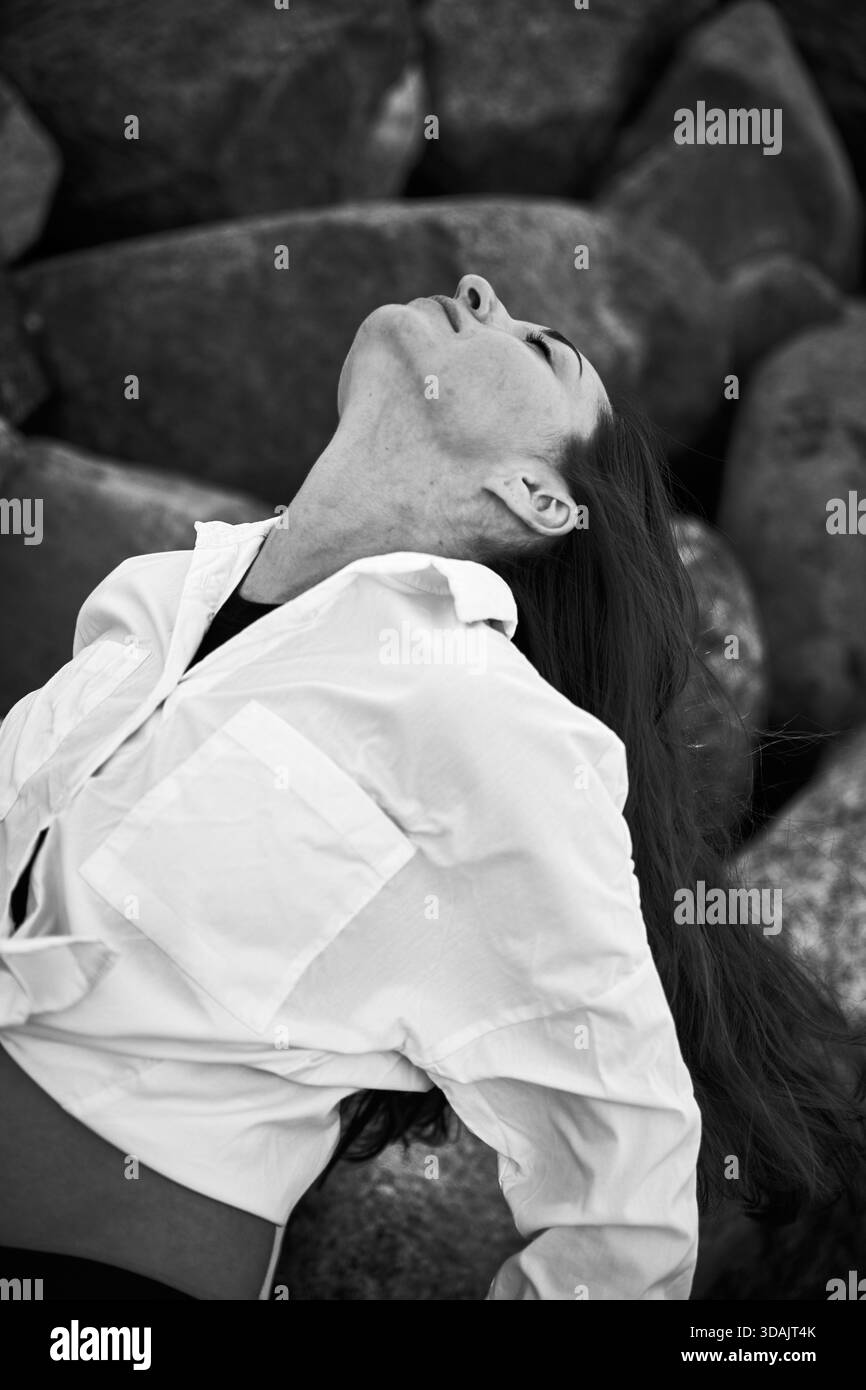 Erwachsene Frau mit langen Haaren lehnt sich mit geschlossenen Augen und entspanntem Ausdruck zurück. Das Schwarzweißbild hebt den Kontrast von weißem Hemd und strukturiertem Stein hervor Stockfoto