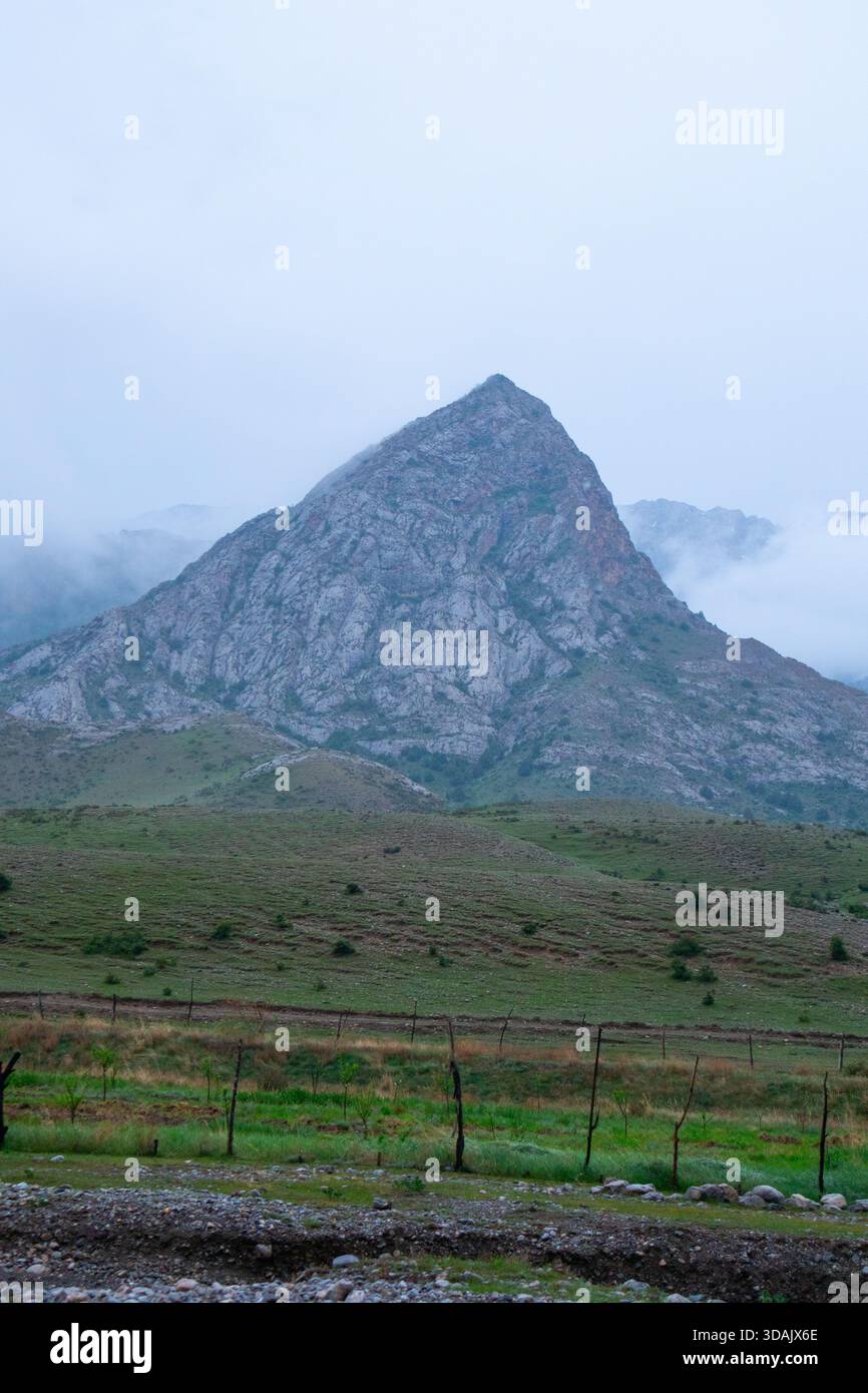Blick auf einen riesigen dreieckigen Felsberg, der über einem grünen grasbewachsenen Tal mit einer Zaunlinie aufragt Stockfoto
