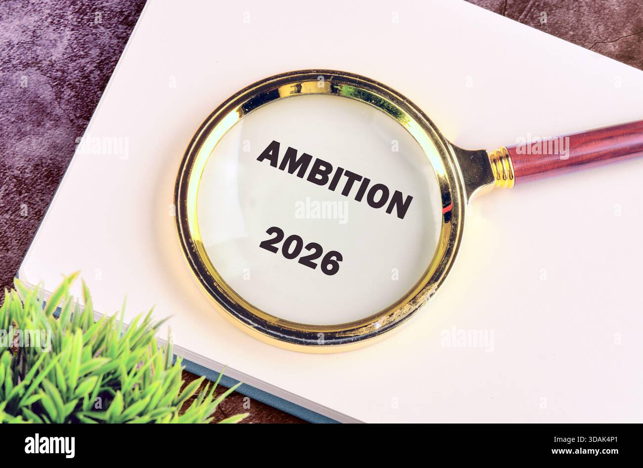 Eine Lupe konzentriert sich auf die Worte AMBITION und 2026, die auf einem weißen Buch neben einer kleinen grünen Pflanze sitzen, was auf sorgfältige Planung und Zukunft hindeutet Stockfoto