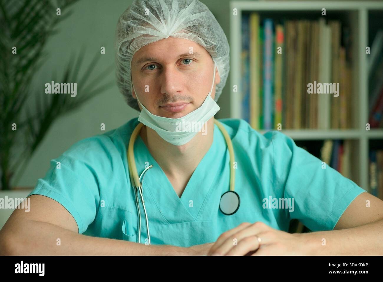 Ein Arzt oder Krankenschwester in medizinischer Uniform mit Maske und chirurgischer Kappe sitzt in seinem Büro. Stethoskop um seinen Hals. Ernsthaft ruhiger Blick in die Kamera. Medizinisches Diagnosekonzept. Stockfoto