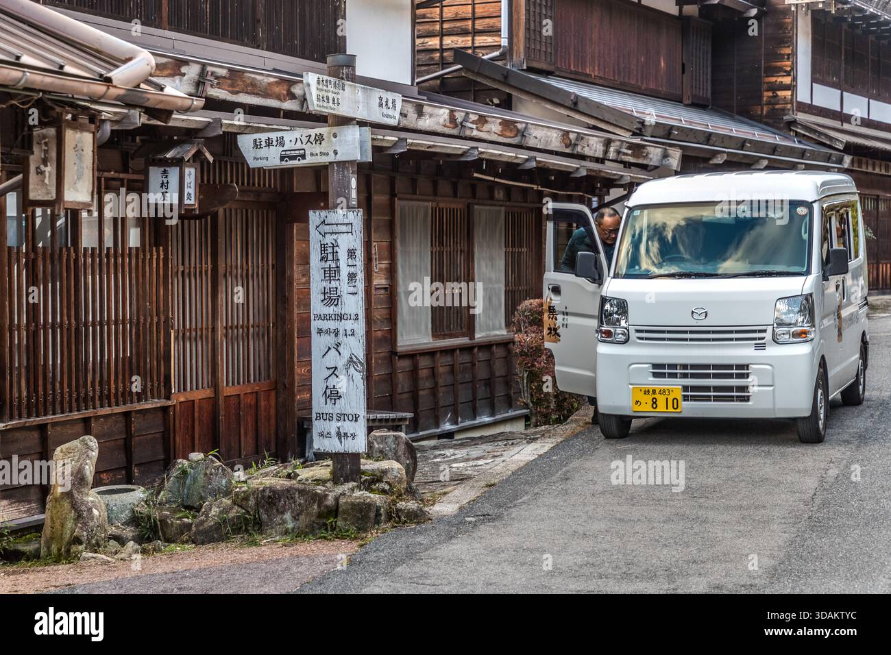 Ein weißer Minibus fährt an einem hölzernen Wegweiser vorbei, der einen Parkplatz und eine Bushaltestelle (バス停) vor der historischen Reihe von Häusern in Tsumago-juku, Nagiso, Präfektur Nagano, Japan anzeigt Stockfoto