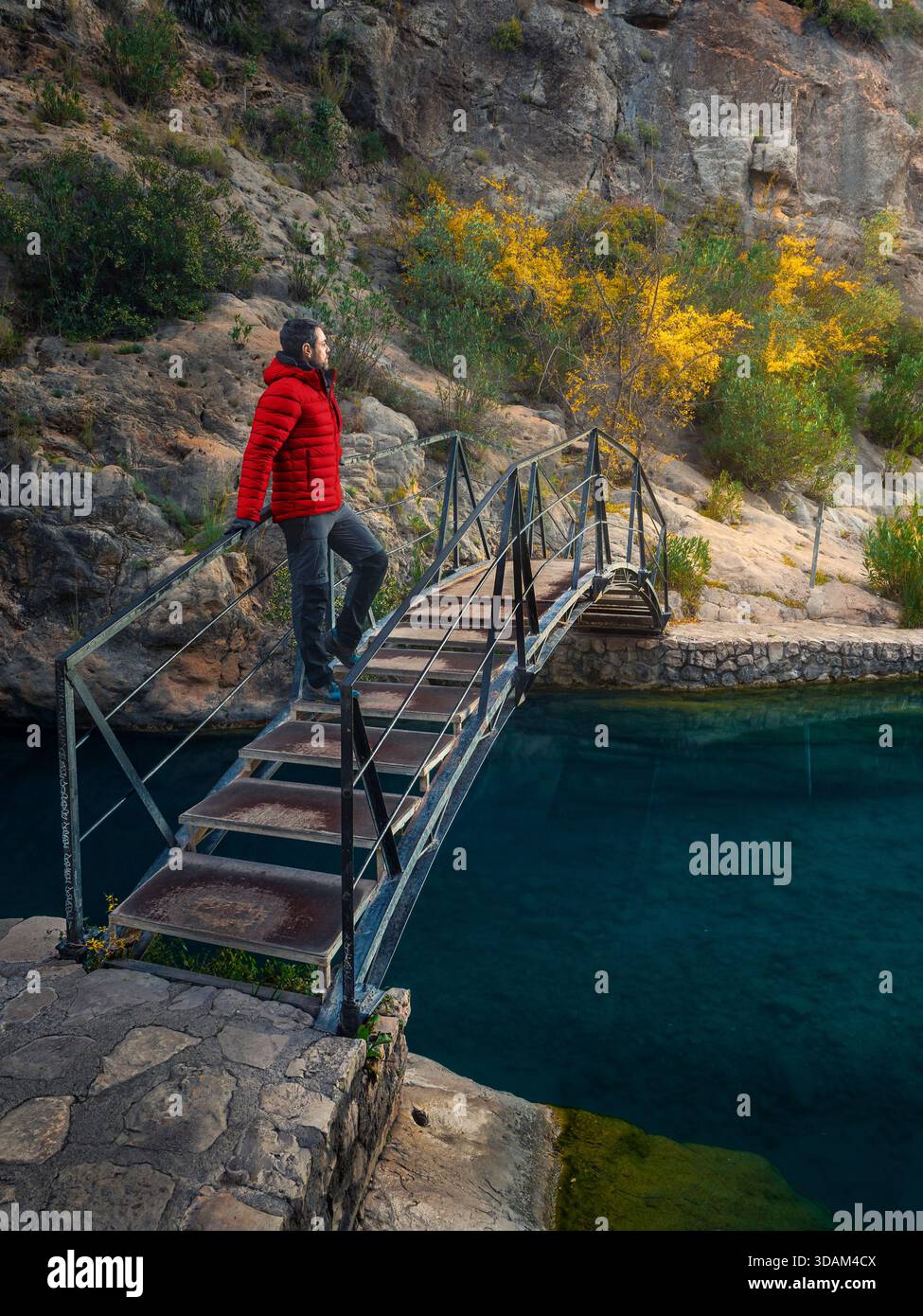 Mann mit rotem Mantel, der auf einer Brücke steht und einen See im Berg beobachtet. Stockfoto