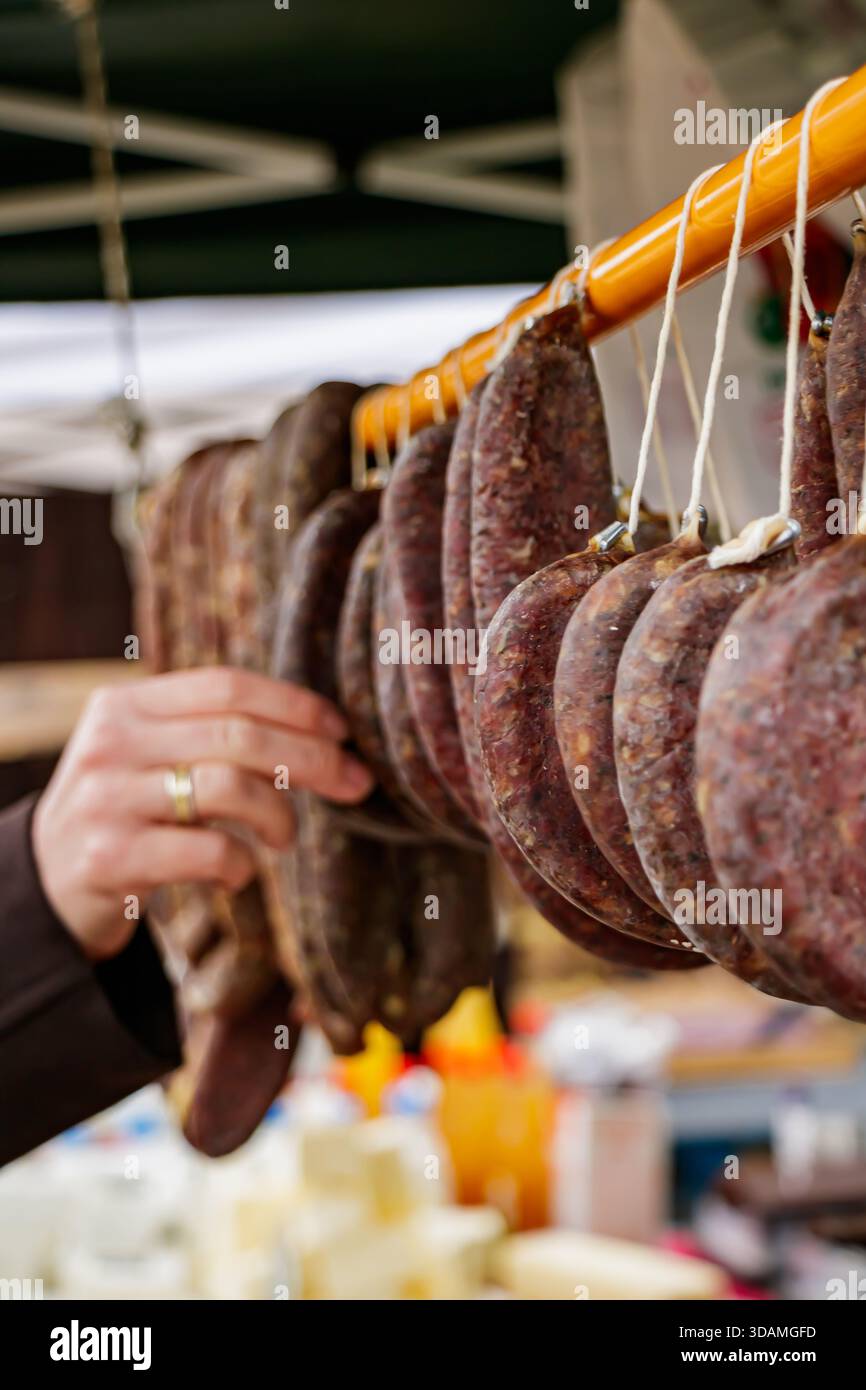 Nahaufnahme der Hand einer Person, die aus Reihen hängender Wurstsorten auf einem Markt im Freien auswählt. Das Bild hebt die authentische handgefertigte Textur von hervor Stockfoto