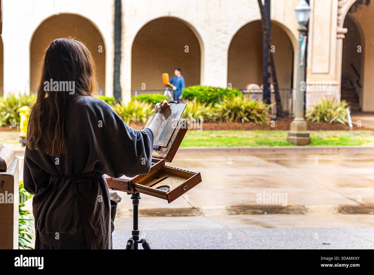 Eine junge Frau malt an einem regnerischen Herbsttag eine Leinwand auf einer Staffelei zwischen den Bögen des Balboa Parks in San Diego, Kalifornien Stockfoto