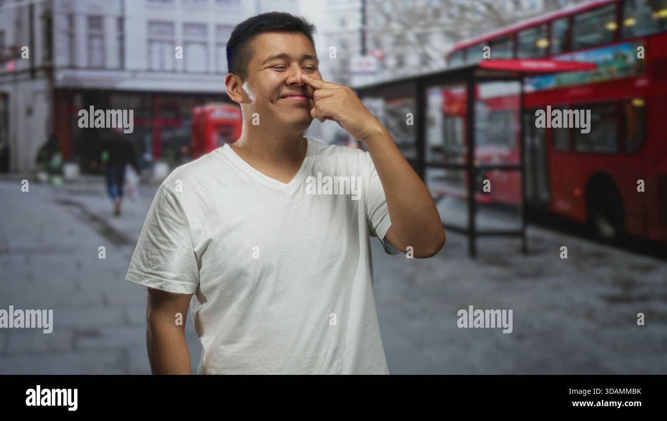 Hispanischer junger Mann im weißen T-Shirt, der seine Nase mit dem Zeigefinger berührt, auf einer Stadtstraße neben rotem Doppeldeckerbus und Telefonbox; Verspieltheit. Stockfoto