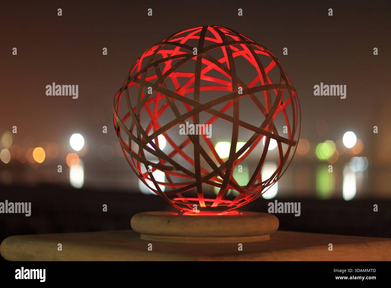 Blick auf eine rot beleuchtete kugelförmige Skulptur mit geometrischen Mustern bei Nacht vor verschwommenen Lichtern der Stadt, Karachi, Sindh, Pakistan. Stockfoto