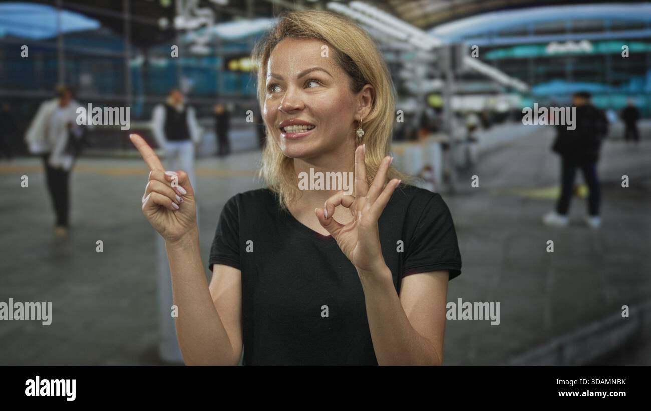 Blonde Frau in schwarzem Hemd macht mit den Fingern am geschäftigen Eingang des Flughafengebäudes ein gutes Zeichen; Optimismus wird gestärkt. Stockfoto