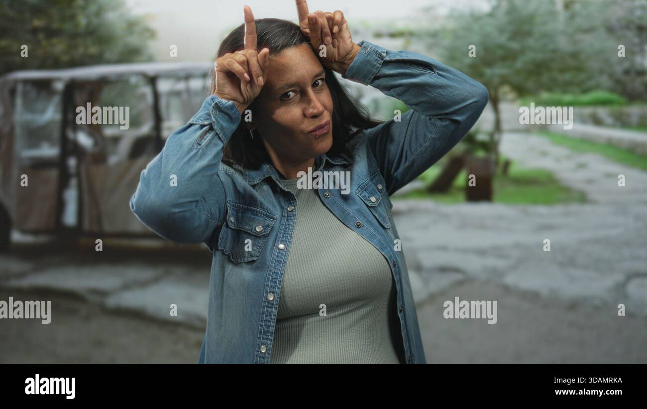 Frau, die Horngesten mit Zeigefingern an den Schläfen macht, Jeansjacke und geripptes Oberteil trägt, auf einer zerrissenen Straße in der Nähe eines geparkten Karren und Baumes steht Stockfoto