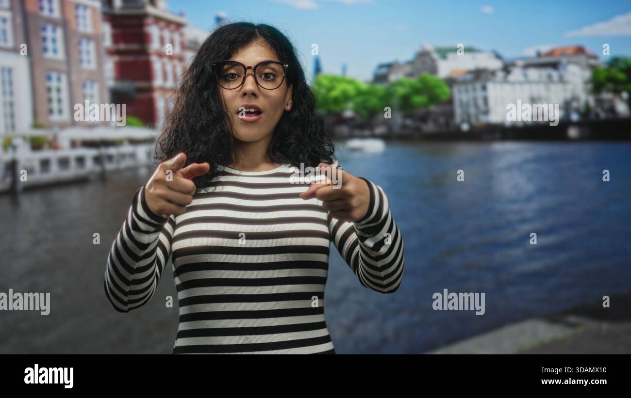 Frau in gestreiftem Oberteil und Brille zeigt mit beiden Händen auf die Straße am amsterdamer Kanal; Vertrauen willkommen. Stockfoto