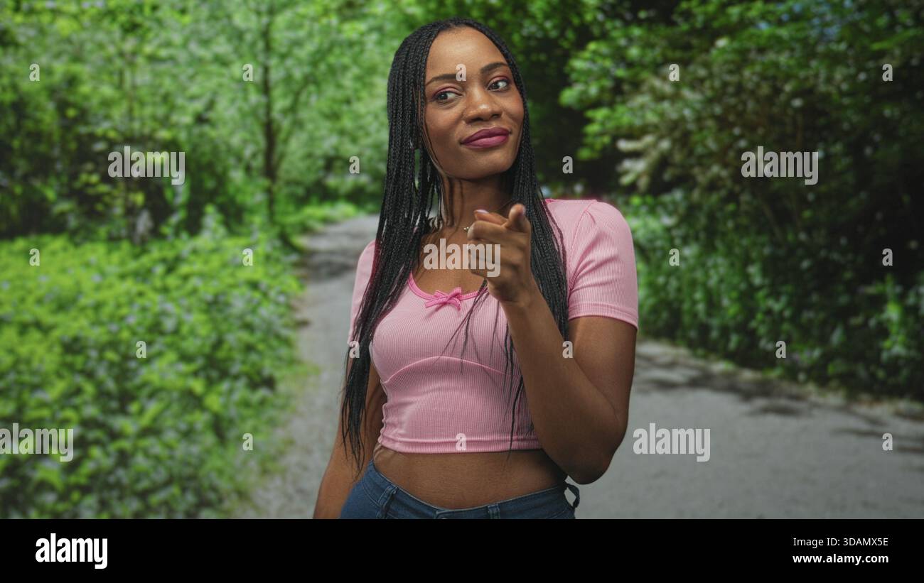 Die lächelnde Frau zeigt den Zeigefinger auf dem Waldweg mit rosa Crop Top und Jeans nach oben; Selbstbewusstsein stärken. Stockfoto