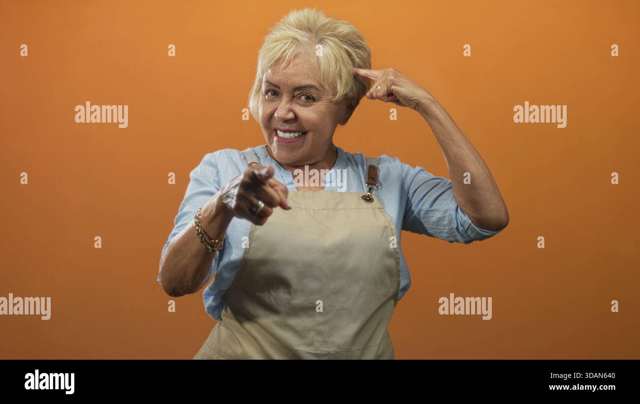 Die ranghöchste hispanische Frau in der Schürze zeigt den Finger auf die Kamera und zeigt den Finger zum Tempel in einem Studio mit orangefarbenem Hintergrund; spielerisches Selbstvertrauen. Stockfoto