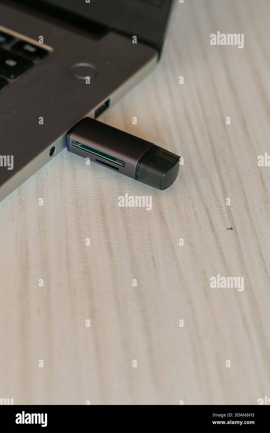 Detaillierte Aufnahme eines dunklen USB-Dongles mit SD-Kartensteckplatz, der in den Laptop-Anschluss gesteckt wurde, auf hellem Holzhintergrund, wodurch die Speichererweiterung hervorgehoben wird. Stockfoto