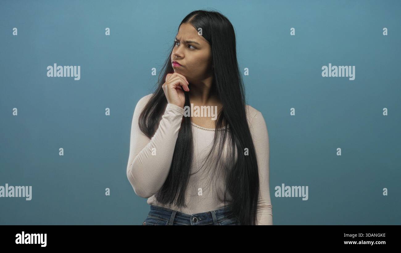 Lateinische Frau posiert mit Hand am Kinn im Studio vor blauer Wand, beugt sich leicht nach vorne und neigt den Kopf; Zweifel. Stockfoto