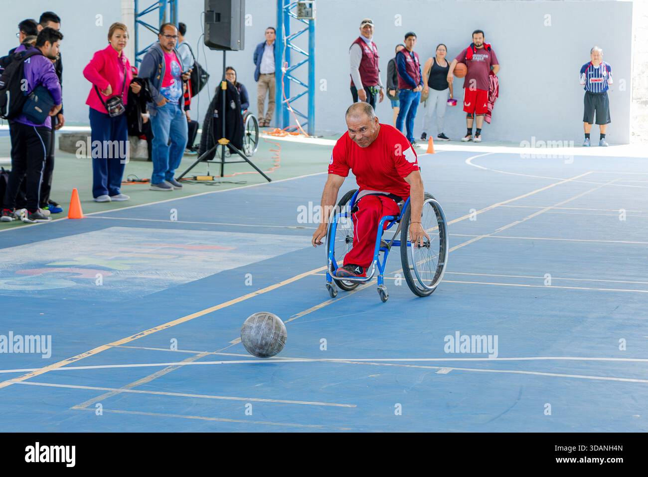 Männlicher Athlet mit Behinderung in einem Rennrollstuhl in Aktion, jagt den Ball in Bodennähe während eines Spiels auf einem Hallenplatz. Stockfoto