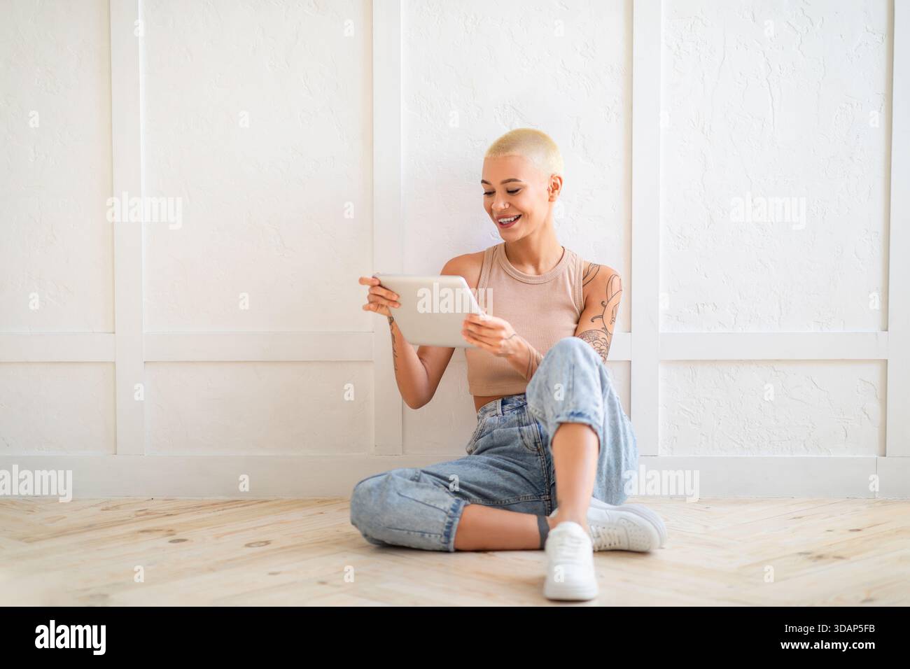 Glückliche junge Frau, die ein digitales Tablet nutzt, im Internet surft oder mit Freunden chattet, auf dem Boden über einer weißen Wand sitzt Stockfoto