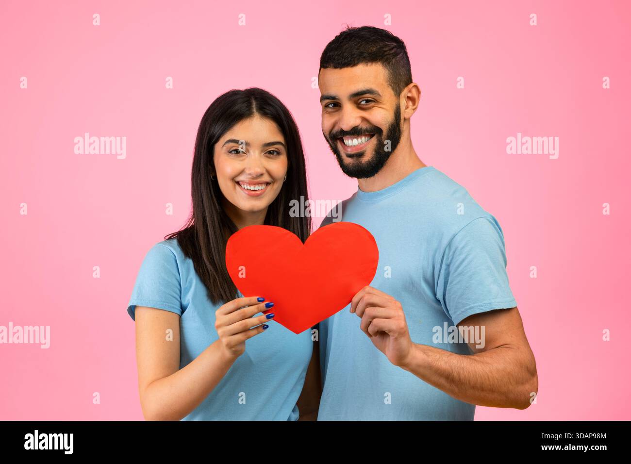 Ein Paar hält fröhlich einen roten Herzausschnitt vor einem rosa Hintergrund Stockfoto