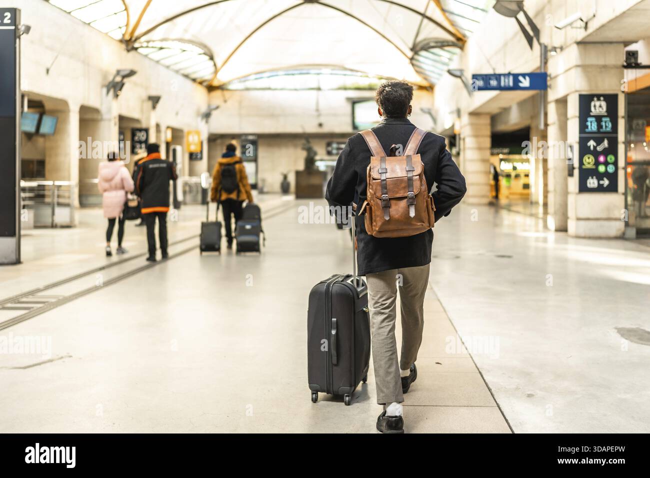 Geschäftsmann, der durch einen modernen Bahnhof geht, einen Koffer zieht und einen Rucksack trägt, hebt Konzepte von Reisen, Pendeln und geschäftsreisen hervor Stockfoto