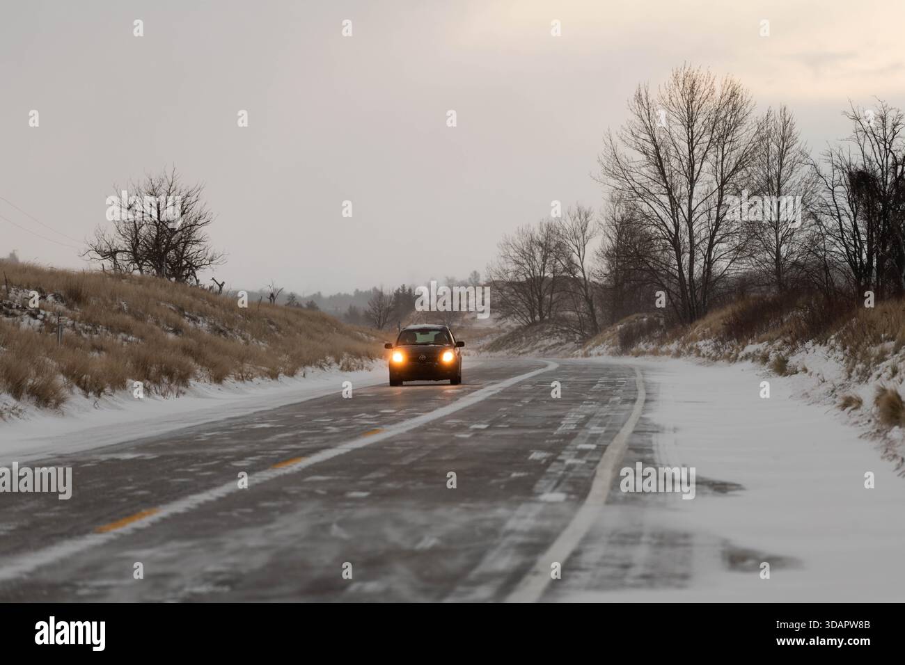 Ein einsames Auto fährt über eine schneebedeckte Straße, umgeben von der ruhigen Schönheit des Ludington State Park im schwindenden Licht des Winters. Stockfoto