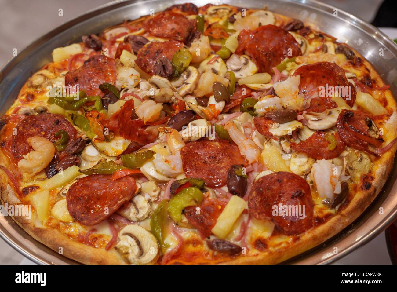 Eine große, gebackene Pizza mit Paprika, Garnelen, Pilzen, Paprika, Oliven, Ananas und Käse stehen auf einem Metalltablett. Stockfoto