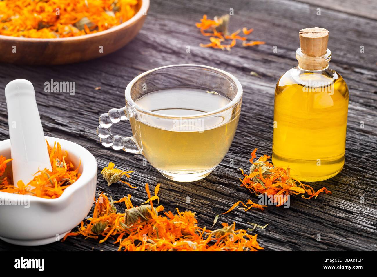 Calendula Tee und ätherisches Öl auf einem rustikalen Tisch, begleitet von getrockneten Blütenblättern und natürlichen Utensilien - Calendula officinalis Stockfoto