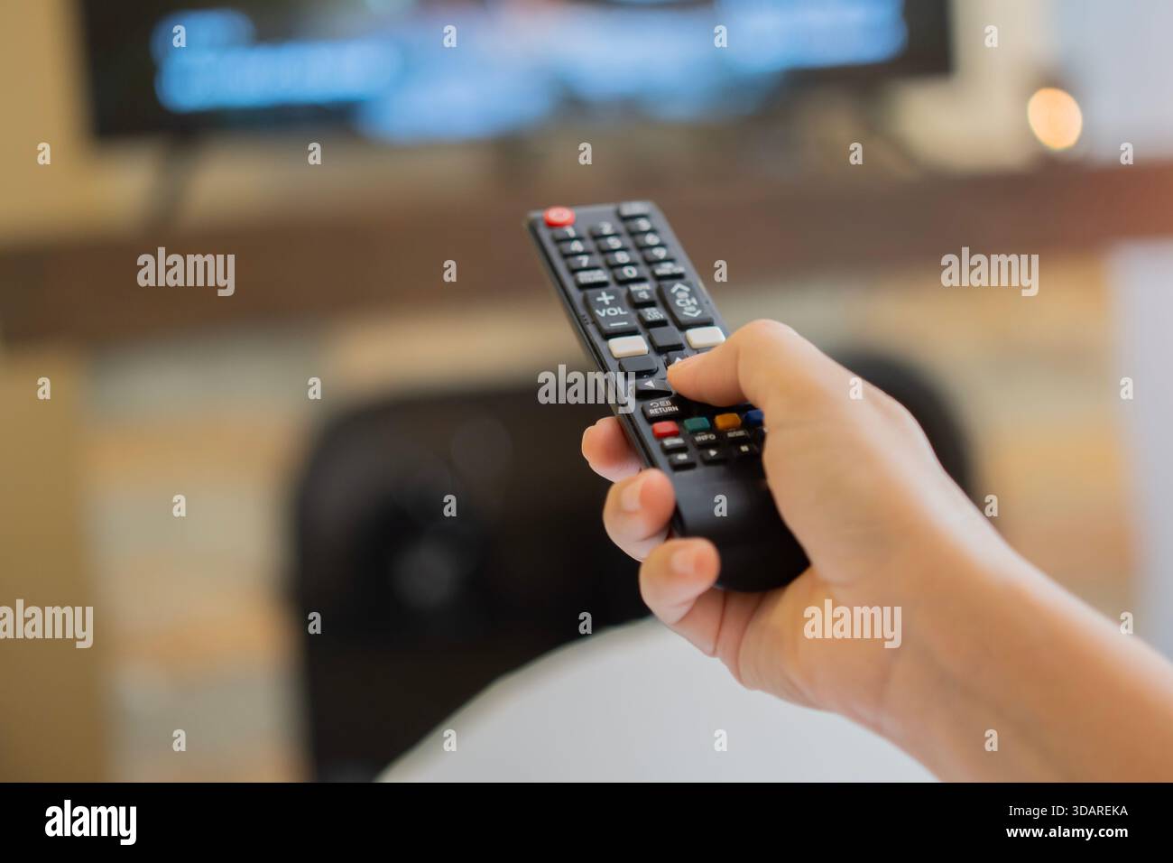 Hand mit tv-Fernbedienung zum Einschalten des Fernsehers zu Hause. Rückansicht einer Frau auf dem Sofa, die den Fernsehsender wechselt. TV-Fernbedienungskonzept. Selektiver Fokus Stockfoto