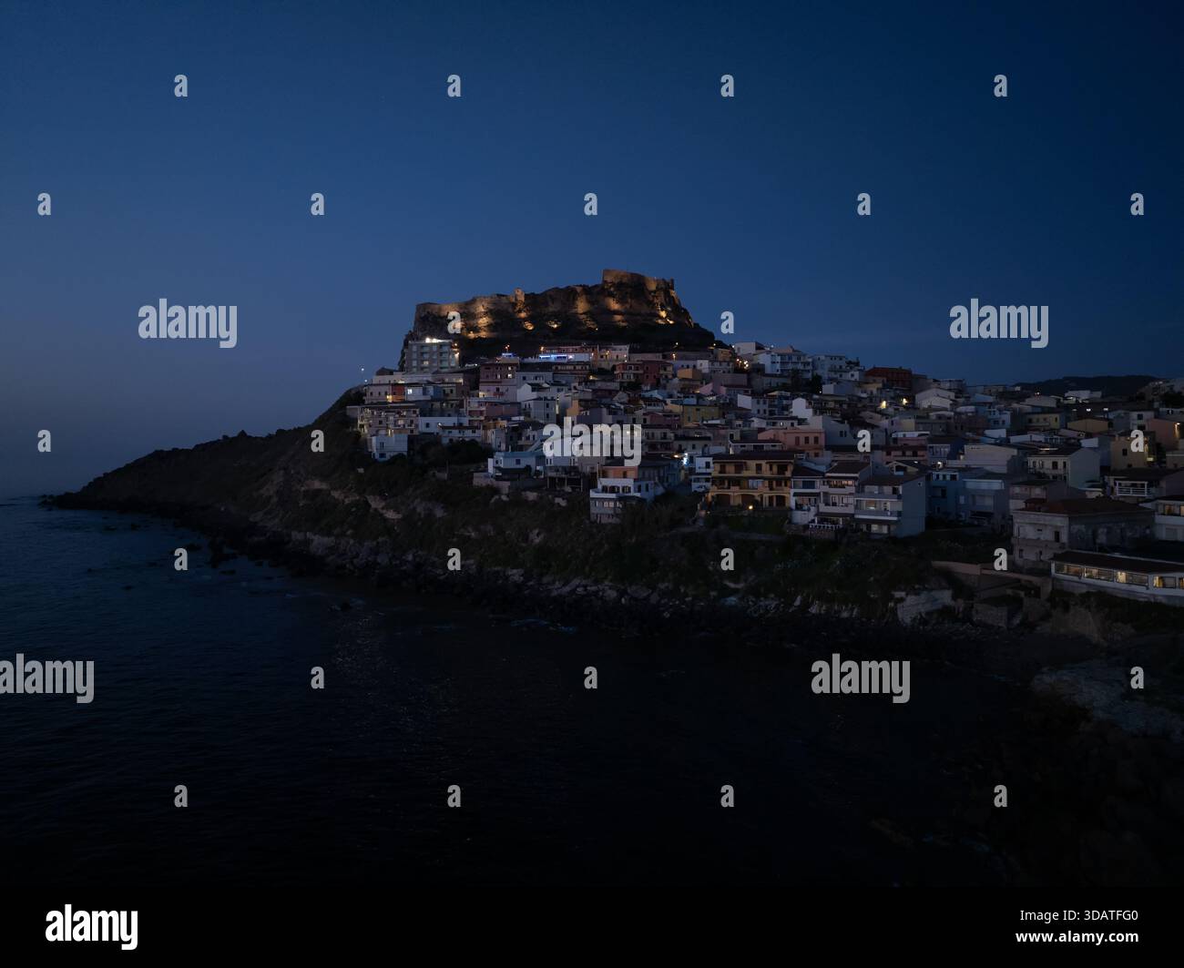 Aus der Vogelperspektive auf das dunkle Wasser trifft auf die zerklüftete Küste unter dem beleuchteten mittelalterlichen Castello dei Doria, Castelsardo, Sardinien, Italien. Stockfoto