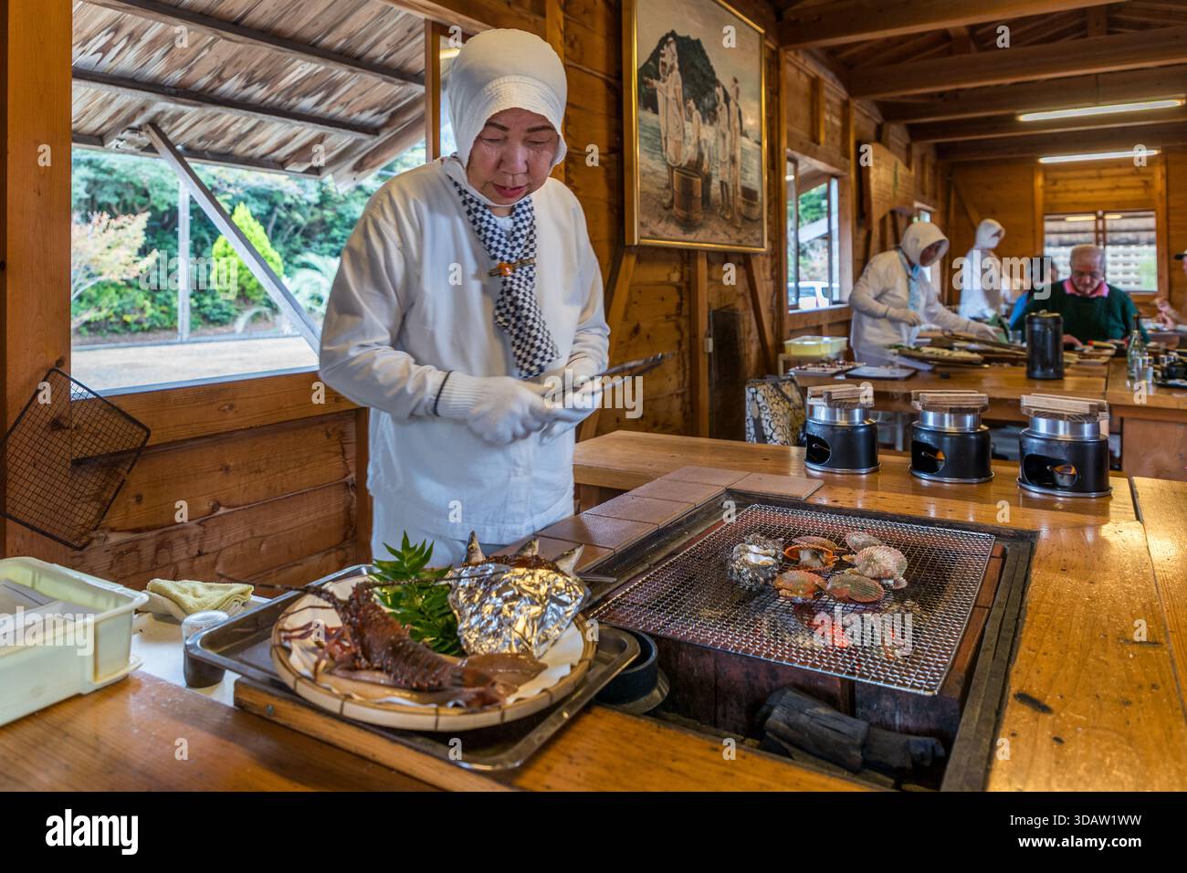 Gäste sitzen um den offenen Grill herum und plaudern direkt mit der Ama-Taucherin Kimiyo Hayashi, während sie frische Meeresfrüchte auf dem Holzkohlegrill zubereitet, in einer Nachbildung einer traditionellen Ama-Hütte, Ise-Bucht, Shima, Präfektur Mie, Japan. Stockfoto