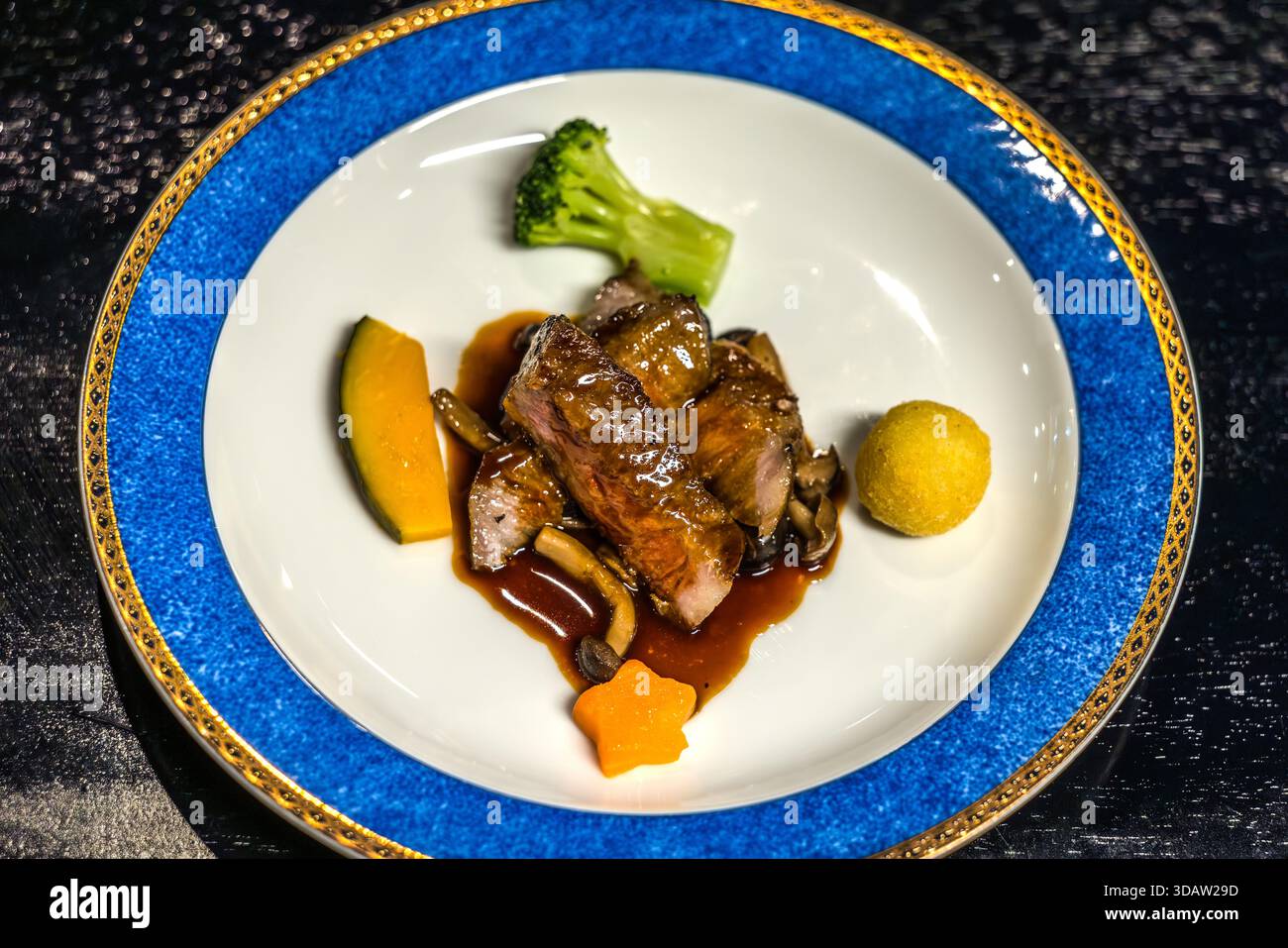 Hauptgericht eines japanischen Kaiseki-Menüs in Toba, Präfektur Mie: Gebratenes Fleisch mit dunkler Reduktionssoße, Shiitake-Pilzen, Brokkoli, Kabocha-Kürbis und Kartoffelkrokette, serviert auf einem weißen Porzellanteller mit blauem Rand und Golddekor. Kaiseki ist eine mehrgängige japanische kulinarische Kunstform, die ihren Ursprung in der Teezeremonie hat und großen Wert auf saisonale Zutaten und ästhetische Präsentation legt. Toba, Mie, Japan Stockfoto