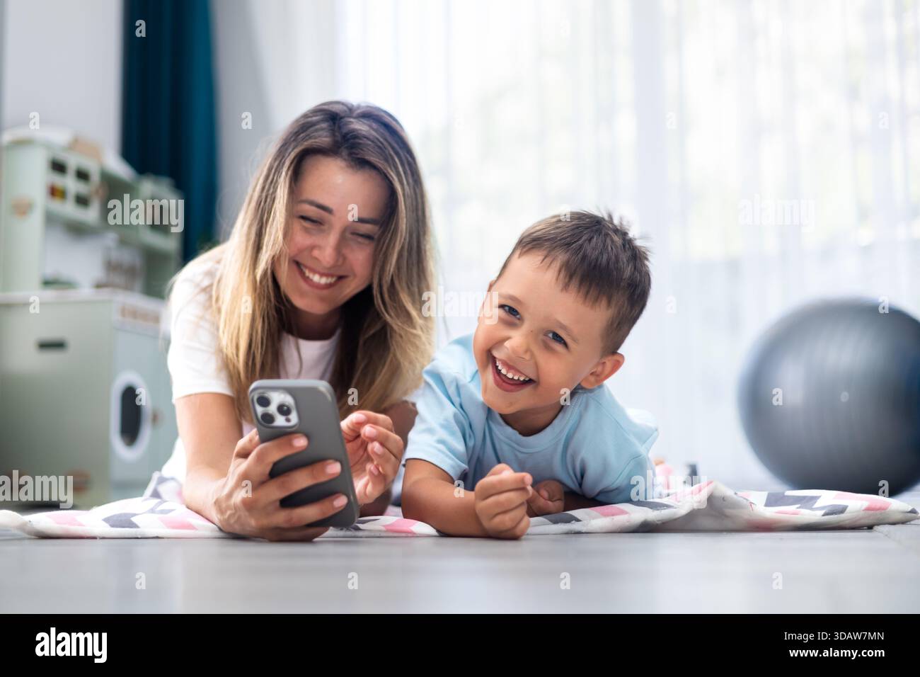Lächelnde Mutter und Sohn liegen auf dem Boden, kleben über ein Smartphone. Der fröhliche Moment fängt Lachen und Konnektivität in einem gemütlichen Wohnzimmer setti ein Stockfoto