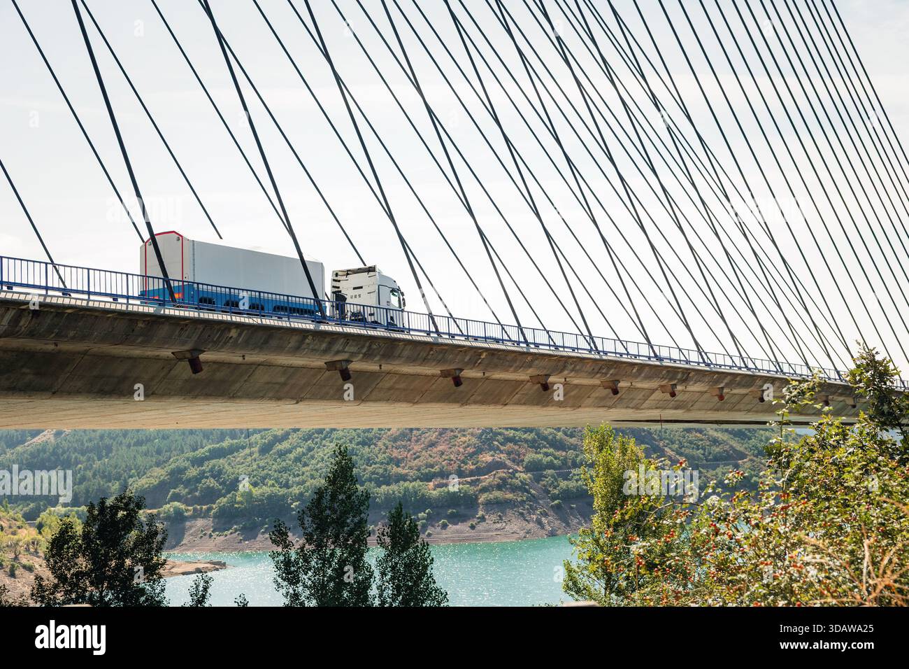 Ein Güterverkehr fährt über eine Seilbrücke unter klarem Himmel, vor dem Hintergrund einer pulsierenden Landschaft mit einem Fluss und grünen Hügeln. Stockfoto