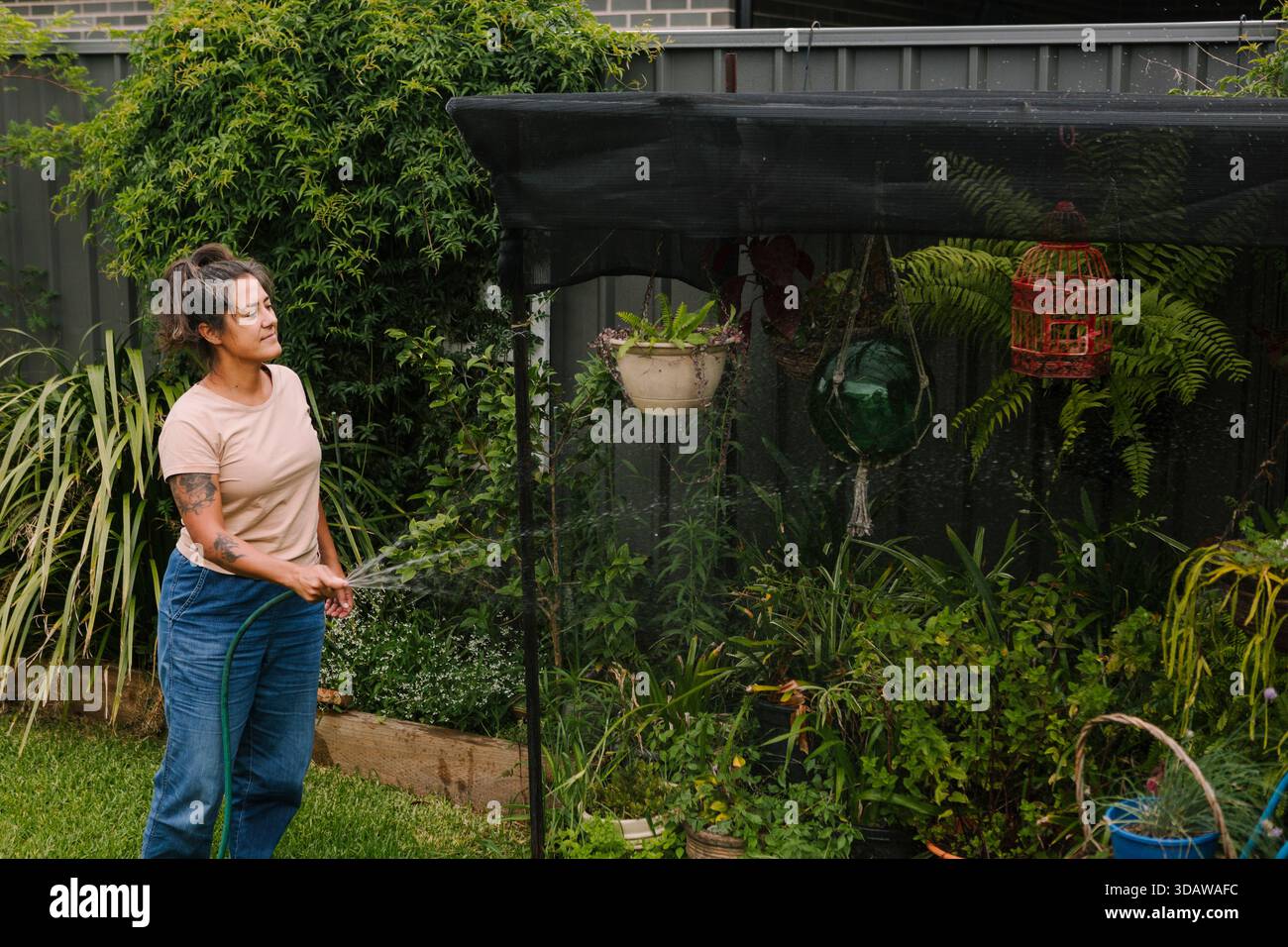 Eine Frau bewässert ihren grünen Garten im australischen Hinterhof, umgeben von einer Vielzahl lebendiger Pflanzen und Blumen. Die Umgebung ist ruhig und erfrischend, Stockfoto