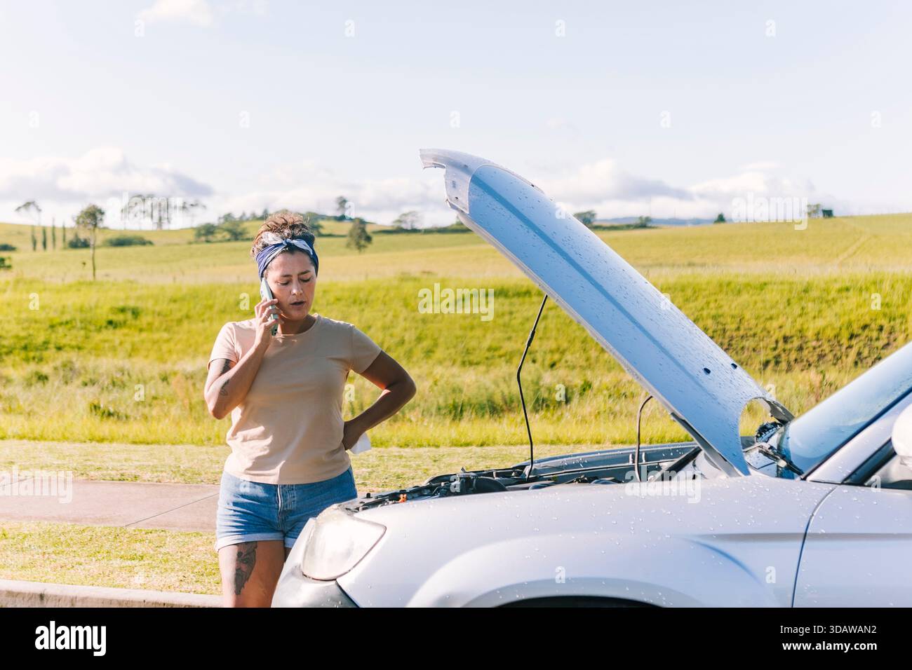 Eine Frau, die neben ihrem kaputten Auto auf einer Landstraße in Australien steht und ein Telefon hält. Die offene Motorhaube und die malerische Landschaft betonen die Isolat Stockfoto