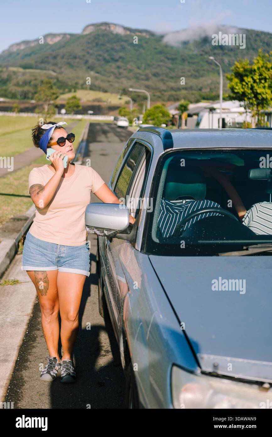 Eine Frau steht an ihrem kaputten Auto an einer australischen Straße und ruft an, um Hilfe zu erhalten. Sie scheint ruhig zu sein und wartet auf Hilfe unter A Stockfoto