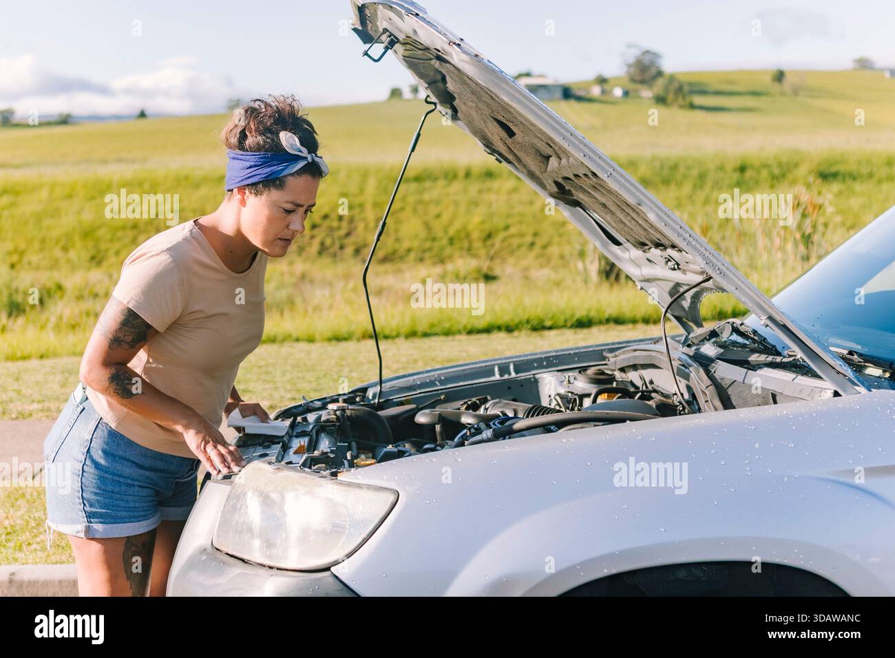 Eine Frau steht neben ihrem kaputten Auto an einem sonnigen Tag in der australischen Landschaft. Sie sieht besorgt aus, als sie den Motor mit der Motorhaube inspiziert Stockfoto