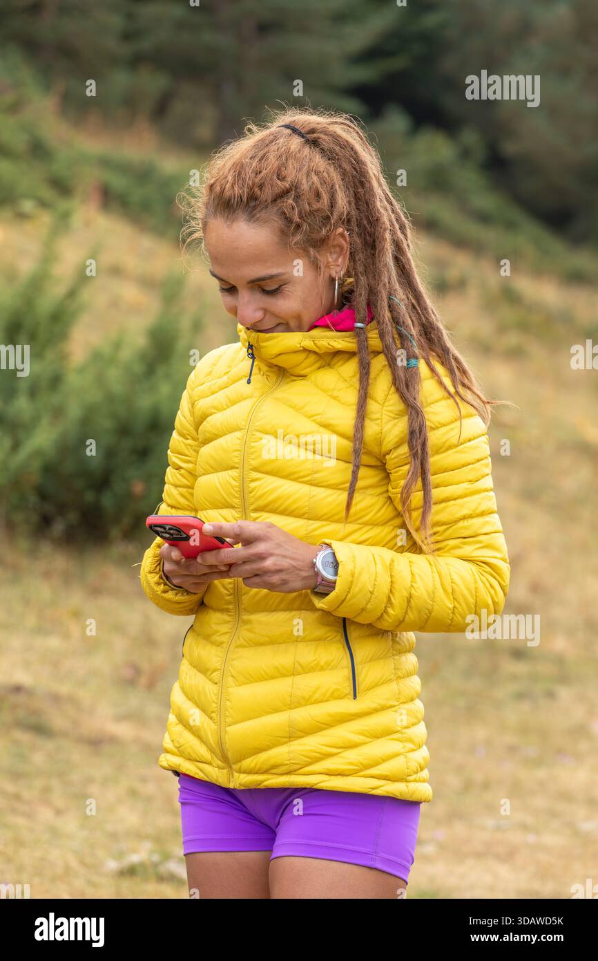 Frau in einer gelben Jacke mit Dreadlocks benutzt ihr Smartphone draußen. Sie genießt einen friedlichen Moment in der Natur, trägt eine Uhr und sieht zufrieden aus Stockfoto