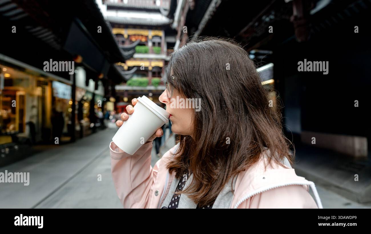 Eine Frau genießt einen Kaffee aus einer Einwegtasse in den lebhaften Straßen von Shanghai, China, umgeben von traditioneller Architektur und geschäftigem Stadtleben Stockfoto