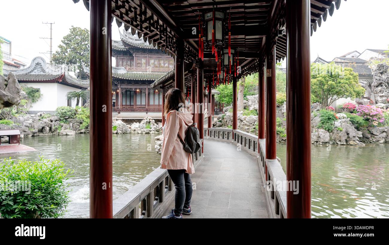 Eine Frau steht auf einem überdachten Gehweg im Yuyuan-Garten in Shanghai, China, und bewundert die malerische Aussicht auf komplizierte Architektur und üppiges Grün. Stockfoto