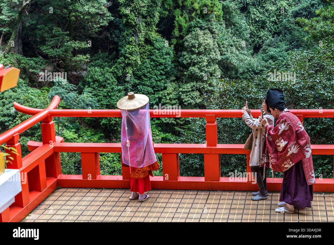 Ein Paar in Heian Kimonos während eines privaten Fotoshootings an den Nachi Falls, Kumano Nachi Taisha. Zwei Personen fotografieren mit ihrem Smartphone eine Person in traditioneller Kleidung auf einer roten Holzterrasse in Nachikatsuura. Präfektur Wakayama, Japan Stockfoto