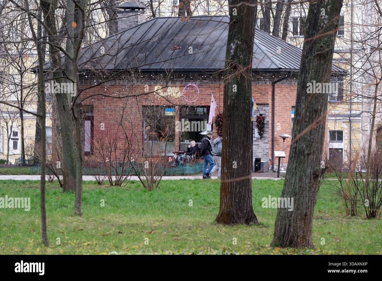 Besucher gehen zu einem charmanten Café, umgeben von hohen Bäumen in einem friedlichen Park. Kiew, Ukraine. Dezember 2025. Stockfoto