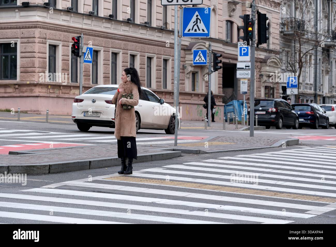 Eine Frau steht geduldig an einem Straßenübergang, während Autos auf einer belebten Straße vorbeifahren. Kiew, Ukraine. Dezember 2025. Stockfoto