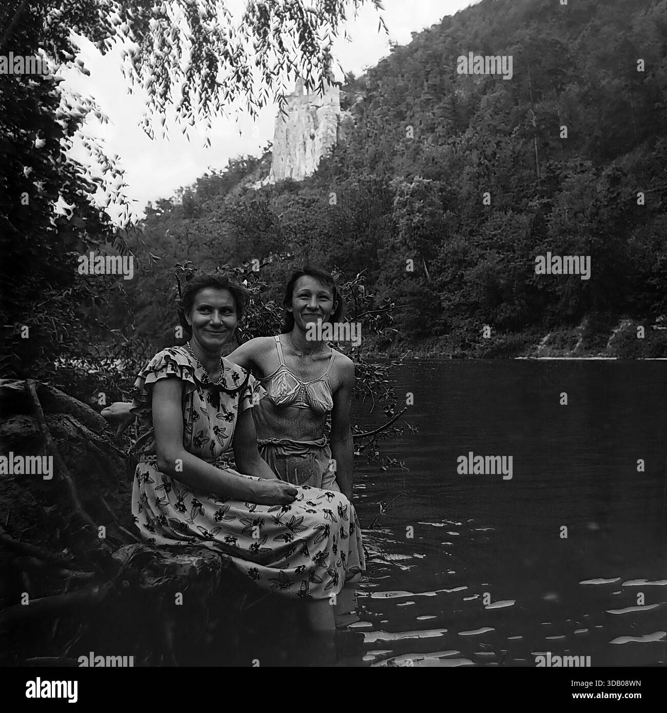 Slavyanogorsk (Svyatogorsk), Ukraine, UdSSR, 1950er Jahre Ein bezauberndes Schwarz-weiß-Archivfoto, das zwei glückliche sowjetische Frauen zeigt, die auf Baumwurzeln am Ufer des Seversky Donets sitzen. Der eine trägt ein Blumenkleid, der andere ein Badeanzug-Oberteil. Im Hintergrund die majestätischen Kreidefelsen und der historische Nikolaikirche über dem Wasser. Dieses Bild zeigt anschaulich weibliche Freundschaft, Sommermode und ruhige Freizeit im friedlichen Donbass. Stockfoto