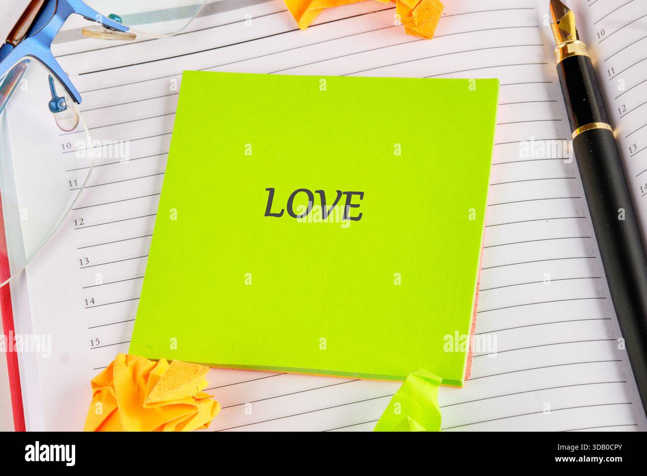 Eine leuchtend grüne Note zeigt das Wort Liebe auf liniertem Notizbuchpapier. Daneben befinden sich ein schwarzer Füllfederhalter, eine Brille und ein verspielter gelber Origam Stockfoto