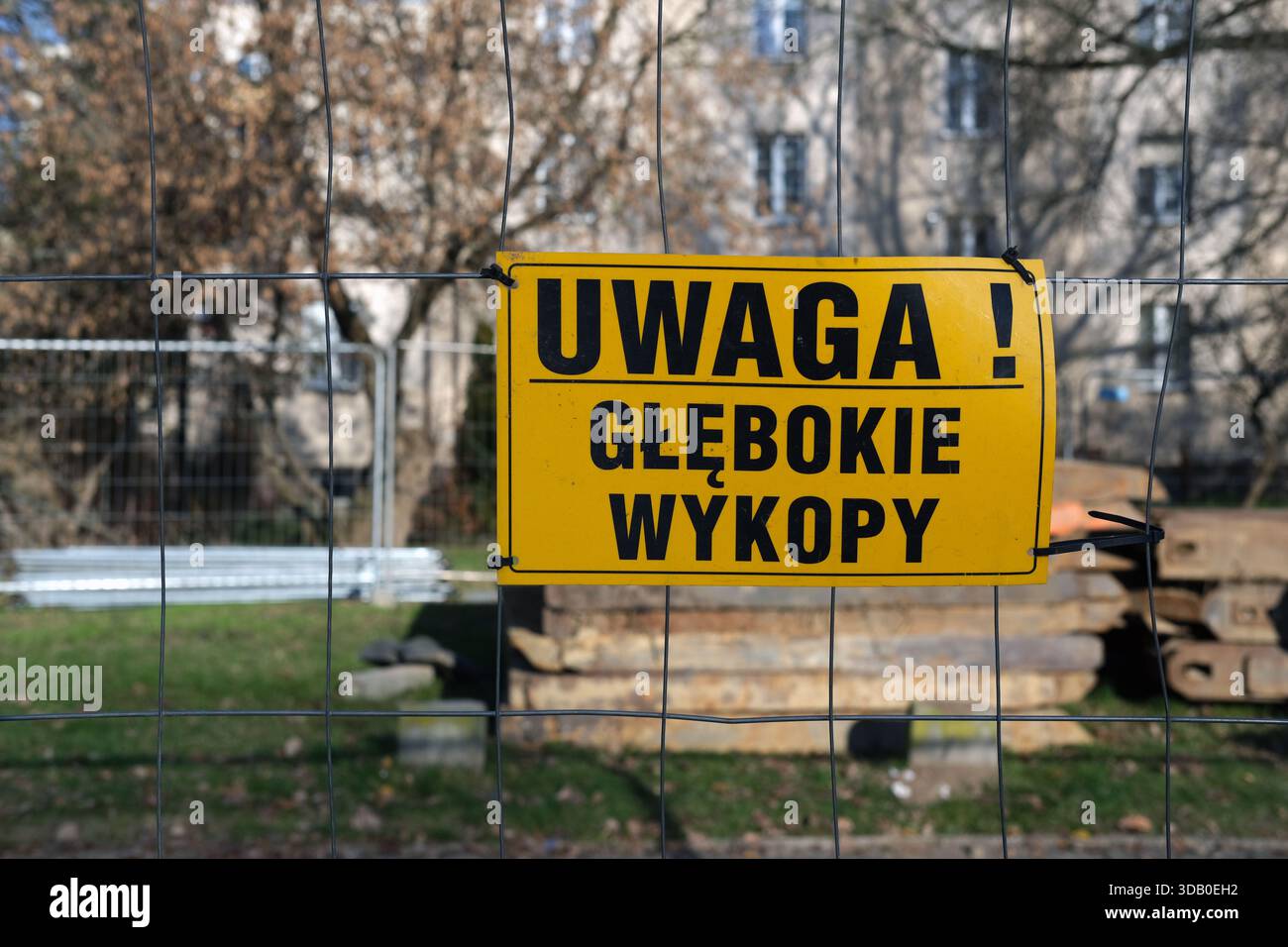 Gelbes Warnschild mit poliertem Text am Metallzaun, Baumaterialien hintereinander gestapelt, Gras im Vordergrund, Tageslicht. Stockfoto