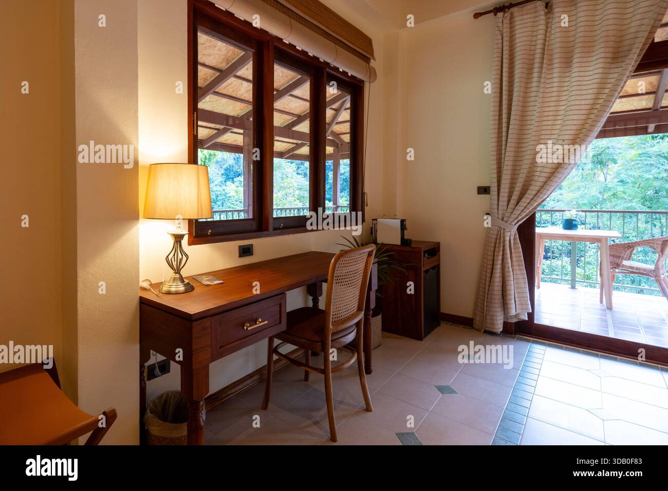 Ein gemütlicher Arbeitsbereich mit Holzschreibtisch und Lampe blickt auf üppiges Grün und schafft eine friedliche Umgebung in Thailand. Perfekt für Entspannung und Inspiration. Stockfoto