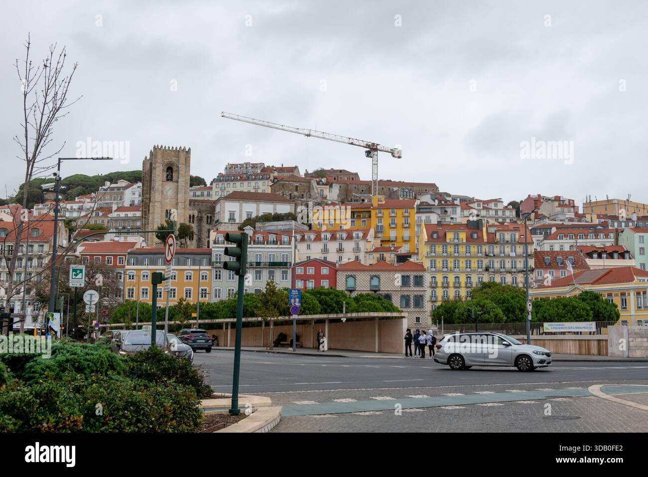 Hinter einer geschäftigen Straße erhebt sich ein lebhafter Hügel mit bunten Gebäuden von Lissabon, eingerahmt von einem historischen Turm und einem Baukran unter einem bewölkten Himmel. Stockfoto